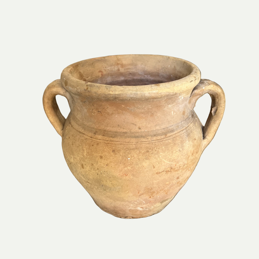 A beige clay pot with two handles, displayed on a wooden surface, with a blurred background featuring wooden elements.