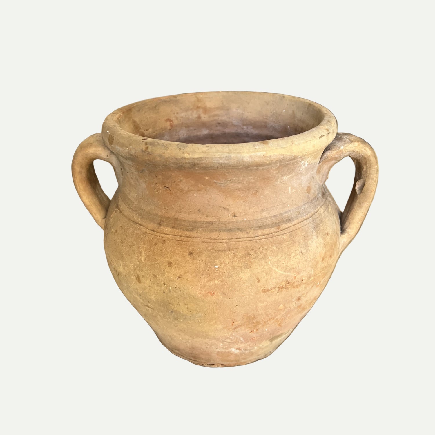 A beige clay pot with two handles, displayed on a wooden surface, with a blurred background featuring wooden elements.