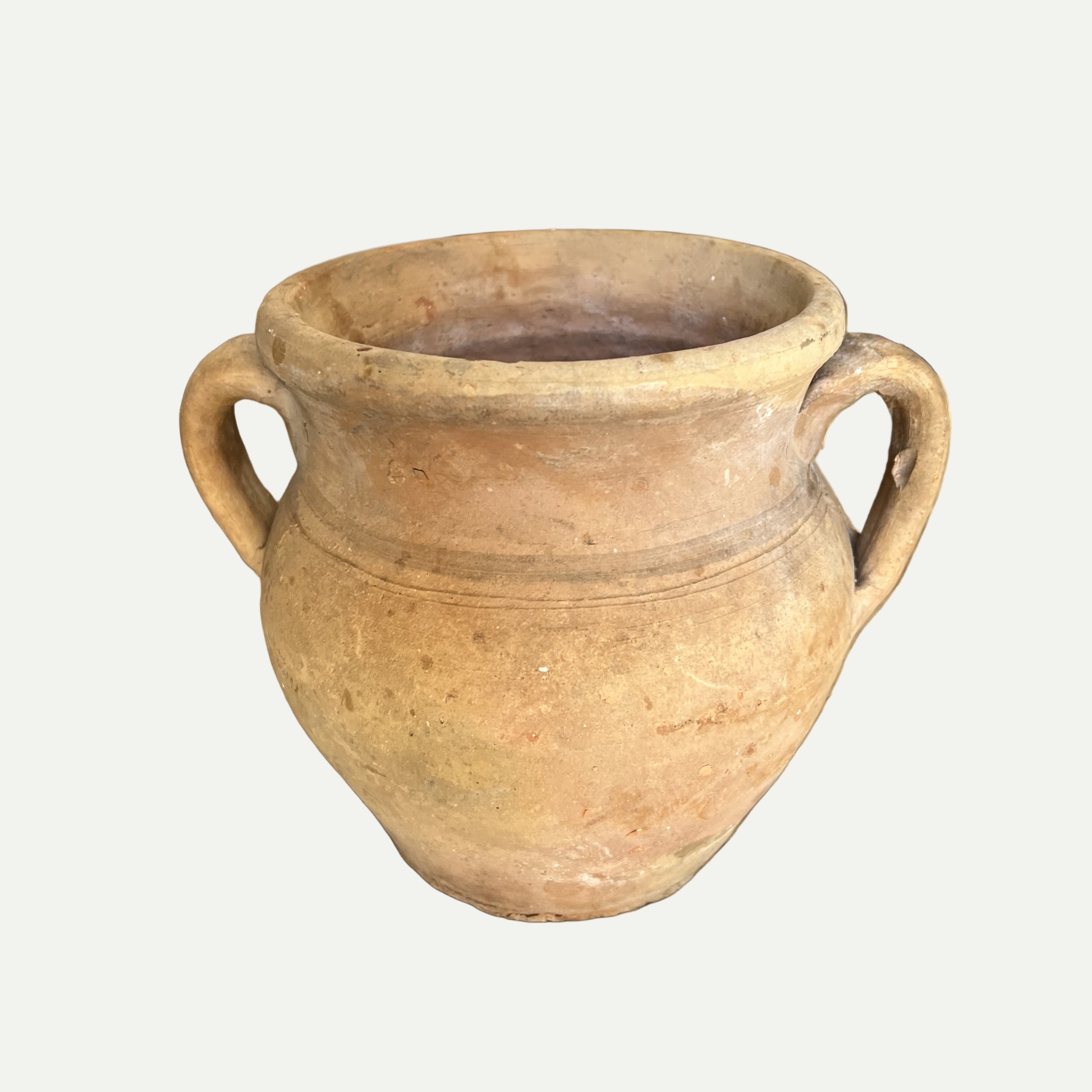 A beige clay pot with two handles, displayed on a wooden surface, with a blurred background featuring wooden elements.