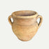 A beige clay pot with two handles, displayed on a wooden surface, with a blurred background featuring wooden elements.