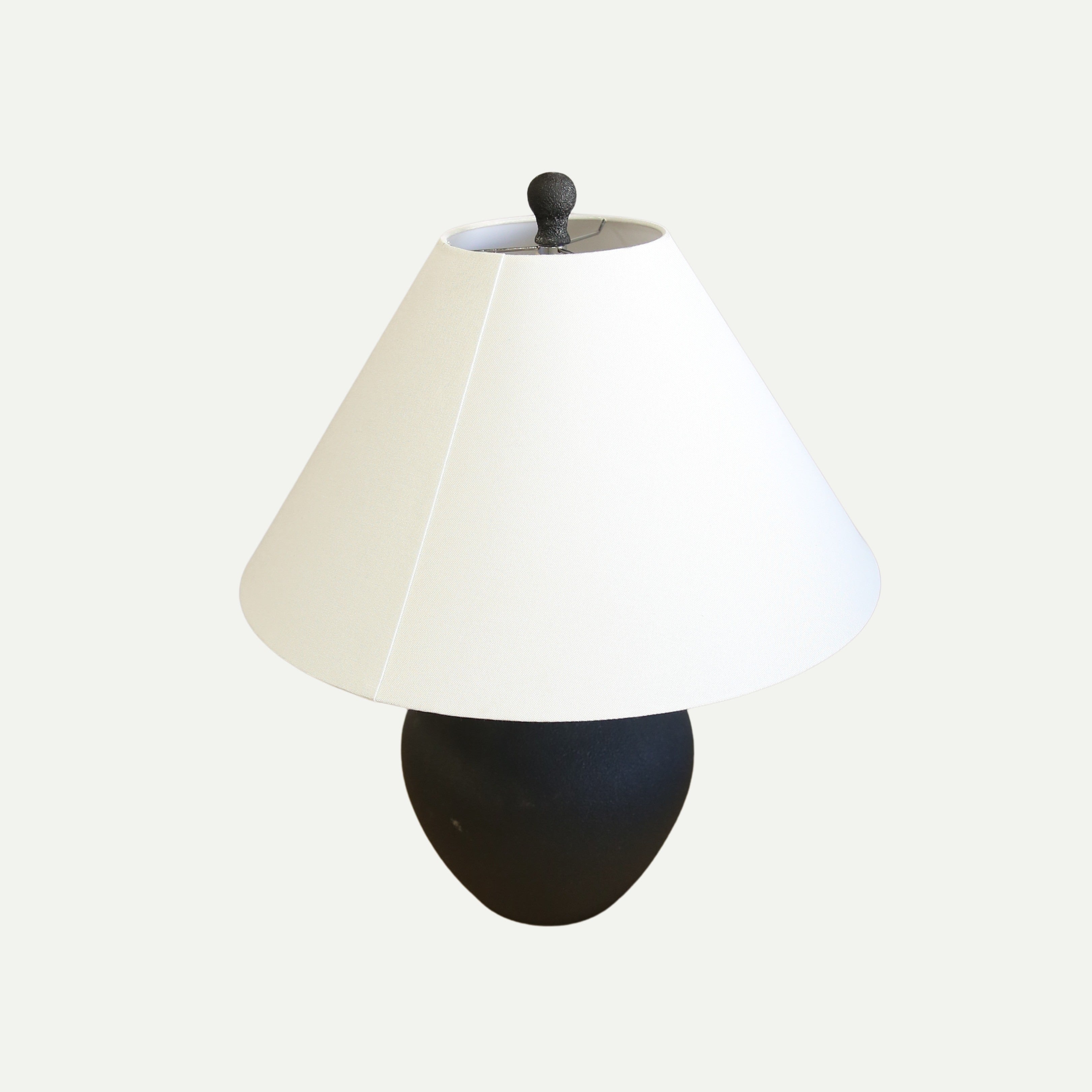 Table lamp with a matte black ceramic base shaped like a vintage amphora and a wide, off-white conical lampshade, set against a plain light background.