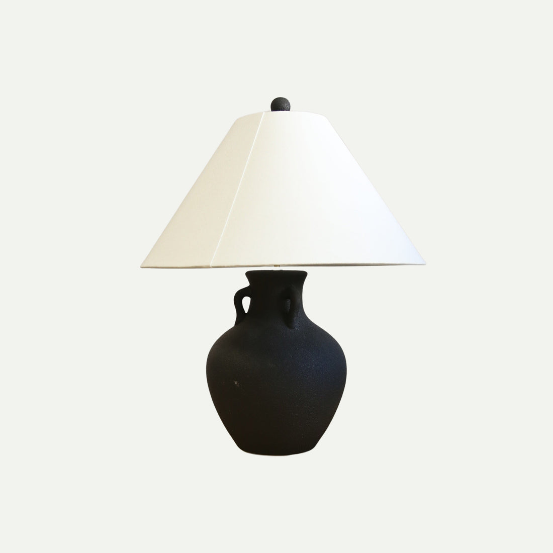 Table lamp with a matte black ceramic base shaped like a vintage amphora and a wide, off-white conical lampshade, set against a plain light background.